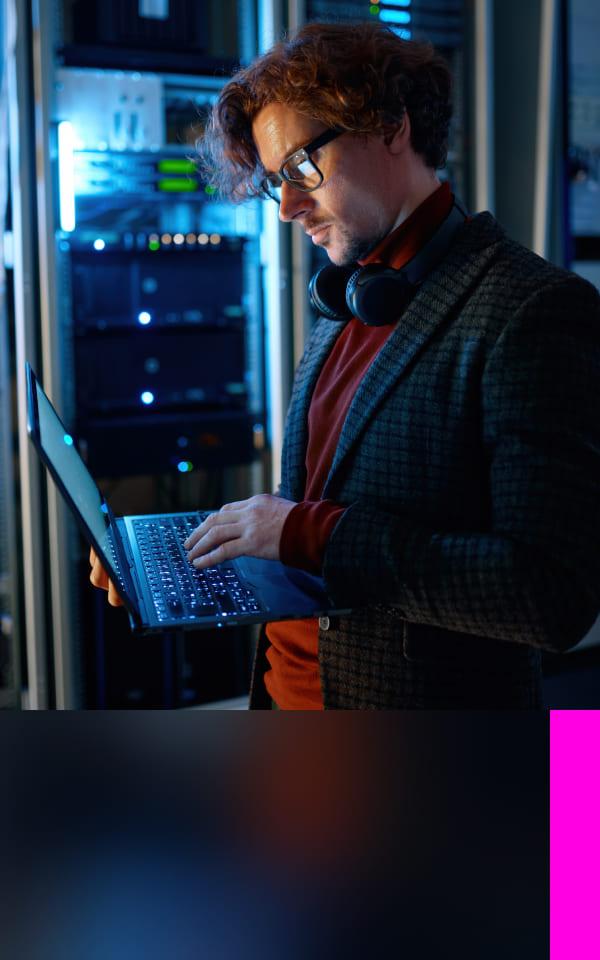 A man with glasses standing behind a server using a laptop to work on their cloud security