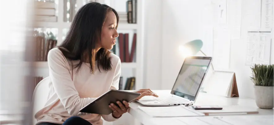 Proteja a sus usuarios que trabajan desde cualquier lugar Mujer trabajando desde casa sosteniendo una tableta y escribiendo en el ordenador